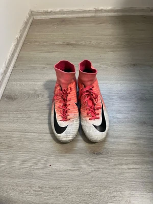 Nike Mercurial rosa fotbollsskor - Säljer ett par Nike Mercurial fotbollsskor i rosa och vitt med svart swoosh. Skorna har högt skaft för extra stöd, snörning och fasta dobbar för spel på gräs. Tillverkade i syntetmaterial som ger bra bollkontroll och passform. Perfekta för dig som vill sticka ut på planen.