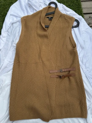 Stickad väst i kamel från H&M - Stickad väst i camelbrun färg från H&M. Modellen är ärmlös med bred krage och två snygga bältesdetaljer i brunt läder på sidan. Grovstickad struktur i mjukt material, perfekt för lager på lager och en chill look.