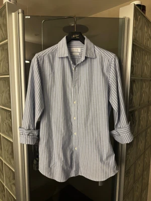 Blåvit randig skjorta - Klassisk blå skjorta med vita ränder från Massimo Dutti. Skjortan har lång ärm, knappar framtill och en stilren krage. Perfekt för en clean och snygg look. .