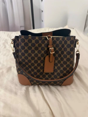 handväska - Köpte för 699kr Snygg brun handväska med klassiskt Triomphe-mönster i guldbrunt. Väskan har detaljer i ljusbrunt läder, guldfärgade metalldelar och justerbar axelrem. Praktisk dragkedja och dekorativ rem framtill. Perfekt för dig som gillar lyxiga accessoarer.