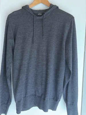 Grå stickad hoodie från Paul Smith - Snygg grå stickad hoodie från Paul Smith i stickad kvalitet. Tröjan har huva med dragsko och ribbade muddar vid ärmslut och nederkant. Perfekt för dig som gillar stilren design och mjuka material.
