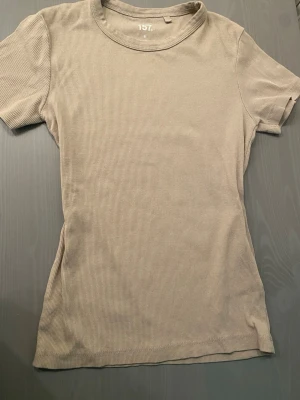 Beige ribbad t-shirt från 157 - En enkel och stilren beige t-shirt från 157 med ribbad struktur. T-shirten har rund halsringning och korta ärmar. Perfekt basplagg som är lätt att matcha med allt i garderoben.