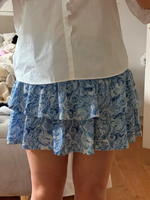 Blå volangkjol med blommigt mönster - Supersöt kort kjol i blått och vitt med blommigt mönster och volanger. Kjolen har en luftig och flowy känsla, perfekt för varma dagar. Kan även användas som en topp. Använd max 3 gånger 