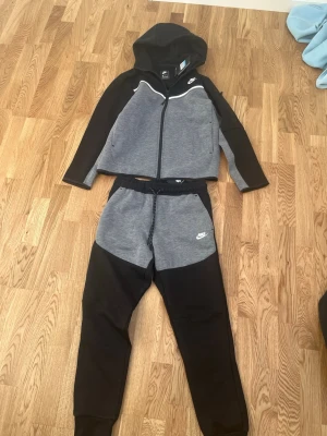 Niketech svar grå  - Nike loungewear set med zip-hoodie och byxor i grått och svart. Hoodien har dragkedja, huva och vita detaljer vid bröstet. Byxorna har dragsko i midjan och ribbade benslut. Båda plaggen har Nike-logga tryckt på framsidan. Perfekt för chill dagar hemma.