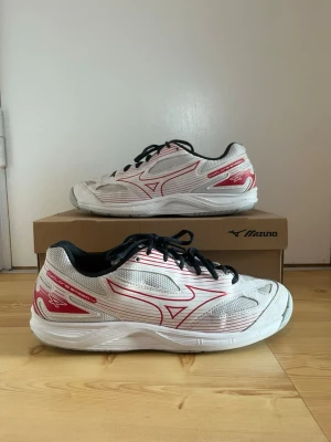 Vita Mizuno sneakers med röda detaljer - Snygga vita sneakers från Mizuno med röda och silverfärgade detaljer samt svart innersula och svarta skosnören. Skorna har en sportig design med meshpaneler för extra ventilation och en bekväm, stötdämpande sula. Perfekta för dig som gillar en clean och modern look.