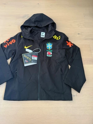 Svart Brasil Nike vindjacka GORE-TEX - Svart vindjacka från Nike med Brasilien CBF-logga, gula och orangea detaljer samt sponsormärken. Jackan har huva, hel dragkedja och är tillverkad i vattentätt GORE-TEX-material. Perfekt för dig som gillar sportig stil och vill visa stöd för Brasilien.