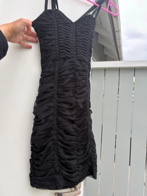 Svart tight klänning med rynk - Säljer en svart figurnära klänning med tunna axelband och snygga rynkade detaljer över hela plagget. Klänningen har en hjärtformad urringning och är sydd i ett stretchigt tyg som formar sig efter kroppen. Perfekt för dig som gillar en trendig och stilren look.H&M stretch bandageklänning.svart festklänning.