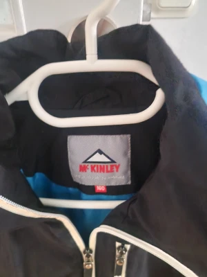 Svart regnjacka McKinley med blå detaljer - Snygg svart regn/vindjacka från McKinley med blå detaljer på axlarna och vit dragkedja. Jackan har en bröstficka med dragkedja och hög krage. Perfekt för regniga dagar och outdoor-äventyr. Tillverkad i lätt och vattenavvisande material.