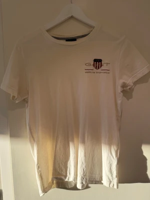 Vit t-shirt från Gant XS - Vit t-shirt från Gant med broderad logga och amerikansk sköld på bröstet. Klassisk rund halsringning och korta ärmar. Tillverkad i mjuk bomull för en bekväm känsla. Perfekt till jeans eller shorts för en clean och enkel stil.