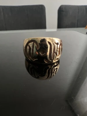 Chrome hearts - Cool bred ring i guldfärgad metall med unik gravyr på framsidan. Ringen har en blank finish och känns riktigt statement på fingret. Perfekt för dig som vill ha något som sticker ut och ger en personlig touch till din stil.