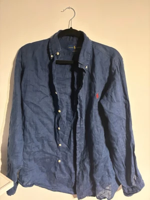 Blå linneskjorta från Ralph Lauren - Klassisk blå skjorta i linne från Ralph Lauren med lång ärm och knappar framtill. Skjortan har button-down krage och den ikoniska röda logotypen broderad på bröstet. Perfekt för en avslappnad men stilren look.