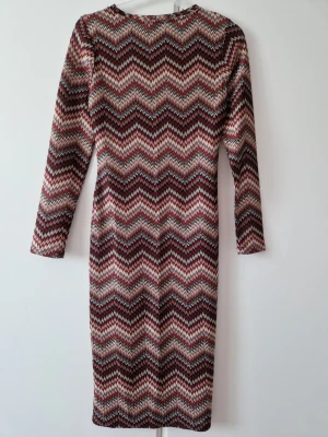 Mönstrad långärmad klänning i zigzag - Snygg långärmad klänning med zigzag-mönster i vinröda, beige och svarta toner. Klänningen har en kroppsnära passform och är i midi-längd. Perfekt för dig som gillar retrovibbar och vill sticka ut med ett coolt mönster.