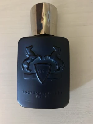 Parfums de Marly Layton - Elegant parfymflaska i matt svart med silverfärgat lock från Parfums de Marly. Flaskan har en stilren, rektangulär form med två upphöjda hästar och en sköld på framsidan. Märkets namn är ingraverat nedtill. Perfekt för dig som gillar exklusiva dofter.