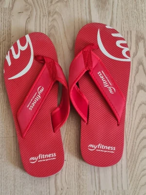 Röda flip-flops från MyFitness - Snygga röda flip-flops från MyFitness med vit logga på sulan. Tillverkade i lätt och tålig syntet med klassisk tårem och räfflad undersida för bättre grepp. Perfekta för stranden eller gymmet.