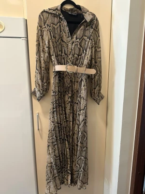 Långklänning med ormmönster från Zara - Snygg långklänning från Zara med beige och svart ormmönster. Klänningen har lång ärm, krage, knappar framtill och ett brett beige bälte i midjan. Perfekt för dig som vill sticka ut med ett trendigt mönster och markerad midja.