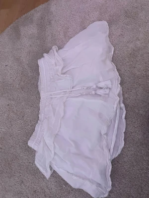 Vit volangkjol med snörning - Söt vit kortkjol med volangkant och snörning i midjan. Kjolen har elastisk midja och tunnt, luftigt tyg som ger en lätt och somrig känsla. Perfekt för dig som gillar en feminin och avslappnad stil.