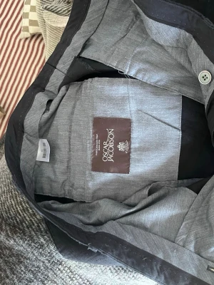 Svart linnekostym från Oscar Jacobson - Snygg svart linne Kostym från Oscar Jacobson med klassisk passform och stilren design. Tillverkad i ullblandning med grått innerfoder och knappar framtill. Perfekt för dig som vill ha en tidlös och elegant look.