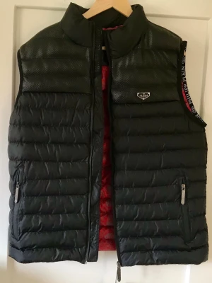 Svart dunväst från Philipp Plein - Snygg svart dunväst från Philipp Plein med glansiga detaljer upptill och logga på bröstet. Västen har två dragkedjefickor och röd insida med mönster. Perfekt för lager-på-lager och streetstyle.