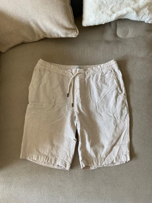 Beige shorts med snörning - Snygga beige shorts med elastisk midja och snörning framtill. Shortsen har sidofickor och två bakfickor med knappar. Perfekta för varma dagar och har en avslappnad passform. Materialet känns lätt och luftigt.