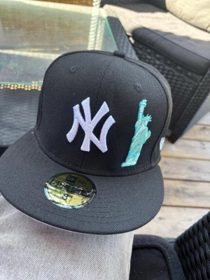 Svart New Era Yankees keps - Svart keps från New Era med New York Yankees-logga i vitt och en broderad frihetsgudinna i turkos på framsidan. Klassisk flat brim-modell med broderad New Era-logga på sidan. Perfekt accessoar för en sportig streetstyle.