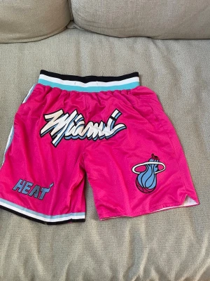 Miami Heat rosa basketshorts - Snygga basketshorts från Miami Heat i stark rosa färg med vit, svart och turkos detalj på midjan och benslut. Stort Miami-tryck framtill och klassisk Heat-logga. Perfekta för sport eller streetwear. Luftigt material som andas.