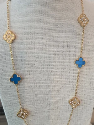 Guldfärgat halsband med blå detaljer - Långt halsband med guldfärgad kedja och fyrklöverformade hängen. Vartannat hänge är blått och vartannat är dekorerat med små gnistrande stenar. Perfekt för att lyfta vilken outfit som helst.