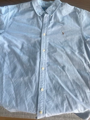 Ljusblå skjorta från Ralph Lauren - Klassisk ljusblå skjorta från Ralph Lauren med knappstängning framtill och broderad logga på bröstet. Skjortan har en tidlös design med krage och långa ärmar, perfekt för en clean och stilren look. Storleken är 20 (XXL) i Juniormått vilket motsvarar S