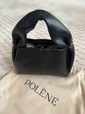 Svart handväska från Polène - Snygg svart handväska från Polène i mjukt läder med unik, skulptural form och bredt handtag. Väskan har guldfärgade detaljer och levereras med original dustbag. Perfekt för dig som gillar stilrena och trendiga accessoarer.