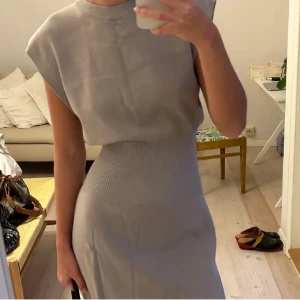 Ljusgrå stickad kortklänning - Superfin ljusgrå stickad kortklänning med markerad midja och ärmlös design. Klänningen har en clean look med rund hals och lätt stretchigt material som sitter snyggt på kroppen. Perfekt för dig som gillar minimalistisk och trendig stil.