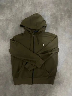 Olivgrön hoodie från Polo Ralph Lauren - Snygg olivgrön hoodie med dragkedja från Polo Ralph Lauren. Klassisk passform med huva, två fickor framtill och den ikoniska vita logon broderad på bröstet. Perfekt för en avslappnad och trendig stil.