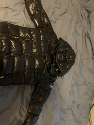 Svart dunjacka från Colmar med huva - Snygg svart dunjacka från Colmar med glansig finish och vadderad design. Jackan har huva, dragkedja framtill och två sidofickor med dragkedja. Perfekt för kalla vinterdagar och riktigt trendig look.