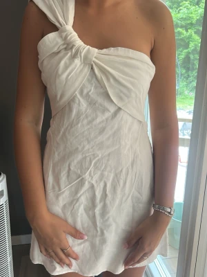 Vit oneshoulder kortklänning - Säljer en vit kortklänning i oneshoulder-modell med snygg knutdetalj på axeln. Klänningen har en enkel och stilren design med lätt vågig nederkant. Perfekt för sommarens alla tillfällen och till student eller skolavslutning!