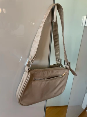 Beige axelremsväska med dragkedja - Stilren beige handväska i avlång modell med bred axelrem och silverfärgade dragkedjor. Väskan har ett ytterfack med dragkedja och är tillverkad i ett slätt syntetmaterial. Perfekt för dig som gillar minimalistisk och enkel design.Har nån fläck som syns på bilderna.