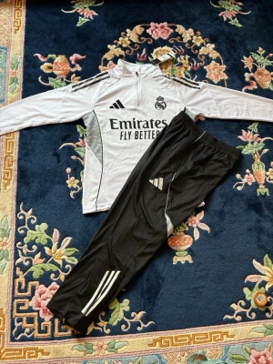 Real Madrid tracksuit - Säljer ett Real Madrid träningsset från Adidas med långärmad vit tröja och svarta byxor. Tröjan har halv dragkedja, klubbmärke och sponsortryck. Byxorna har vita detaljer och Adidas-logga. Perfekt för fotbollsträning eller chill.