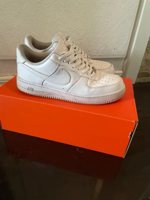 Nike Air Force 1 vita sneakers - Klassiska vita Nike Air Force 1 sneakers i lågt utförande. Skorna har en stilren design med perforerad tå, vit sula och ikoniska swoosh-loggan på sidan. Tillverkade i läder med vita skosnören och diskreta detaljer. Perfekta för en clean och tidlös look.