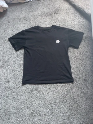 Svart Moncler t-shirt i bomull - Säljer en svart t-shirt från Moncler med klassisk rund hals och korta ärmar. T-shirten har Monclers ikoniska logga broderad på bröstet och är tillverkad i mjuk bomull för en skön känsla. Perfekt till jeans eller shorts för en clean look. Obs ej äkta använd en gång