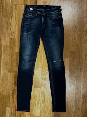Mörkblå jeans från Replay - Storlek: W27 / L32 - Trendiga jeans från Replay i Hyperflex-material som ger en flexibel och följsam passform. Klassisk mörkblå färg som passar till det mesta! Modell: New Luz. Storlek: W27 / L32. Material: 88% Bomull, 8% Polyester och 4% Elastan. Knäpps med knapp och dragkedja. Två framfickor och två bakfickor. Tillverkade i Italien. Säljs i begagnat skick, hål finns på ena knät (se bilder för detaljer) Kika gärna in på mina andra annonser, jag säljer just nu flera klädesplagg! Hör av dig vid frågor eller intresse!