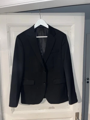 Svart klassisk kavaj från dressman  - Stilren svart kavaj med två knappar framtill och klassiska slag. Kavajen har två fickor med lock och en bröstficka. Fodrad insida och smal passform som ger en snygg siluett. Perfekt för dig som vill ha en tidlös look.