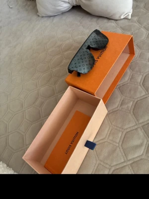 Svarta Louis Vuitton solglasögon - Säljer ett par svarta solglasögon från Louis Vuitton med rektangulär sköldform och ikoniskt LV-mönster på glaset. Snygg och modern design med breda bågar och mörka linser. Perfekt accessoar för dig som vill sticka ut. bra skick 7/10 här ingår boxen och papprerna hittar tyvärr inte påsen dom brukar va i! o dom är i E modellen! 