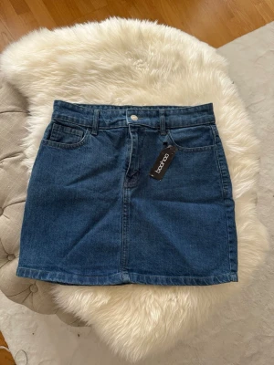 Mörkblå jeanskjol från Boohoo - Säljer en klassisk mörkblå jeanskjol från Boohoo. Kjolen har fem fickor, bälteshällor och stängs med knapp och dragkedja framtill. Perfekt basplagg med rak passform och snygg denimlook.