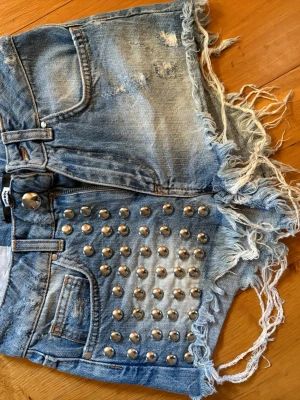 Blå jeansshorts Bikbok XS - Säljer ett par blå jeansshorts med slitna detaljer, fransiga kanter och silverfärgade nitar framtill. Shortsen har klassisk femficksdesign och bälteshällor. Perfekta för dig som vill ha en edgy och trendig look.