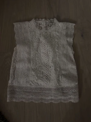 Vit spetsblus med volangkant - Superfin vit blus i spets med vackra volangkanter och hög hals. Blusen är ärmlös och har detaljerad spets med blommigt mönster och dekorativa kanter nertill och vid ärmöppningarna. Perfekt för dig som gillar romantisk och feminin stil.