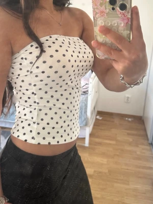 Vit prickig tubtopp - Snygg vit tubtopp med svarta prickar. Toppen är axelbandslös och har en tight passform som sitter bekvämt mot kroppen. Perfekt att styla med jeans eller kjol för en trendig look.