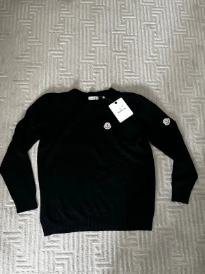 Svart stickad tröja från Moncler - Svart stickad tröja från Moncler med rund halsringning och diskret Moncler-logga på bröstet och ärmen. Tröjan har ribbade muddar vid ärmslut och nederkant. Perfekt för dig som gillar stilrena och exklusiva plagg.