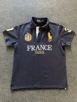 Marinblå pikétröja från Ralph Lauren - Snygg marinblå pikétröja från Ralph Lauren med FRANCE PARIS-tryck och broderade detaljer i vitt och guld. Klassisk krage, kortärmad och med knappar framtill. 