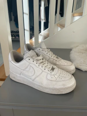Nike Air Force 1  - Klassiska Nike Air Force 1 sneakers i helvitt läder med perforerad tåbox och ikonisk swoosh på sidorna. Skorna har en chunky sula och lågt skaft, perfekt för en clean och tidlös streetstil.