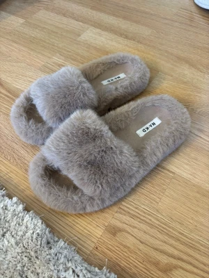 Beige fluffiga tofflor från NA-KD - Supermysiga beige tofflor från NA-KD med fluffig pälsimitation och öppen tå. Perfekta att glida runt i hemma, med mjuk ovandel och svart räfflad sula för extra grepp. Snygg och trendig design som känns lyxig och cozy. 