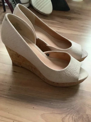 Söta vita kilklackar  - Säljer ett par beiga kilklackar från H&M med öppen tå och strukturmönstrad ovandel. Skorna har en snygg korkklack som ger extra höjd och somrig känsla. Perfekta för dig som vill ha både stil och komfort. Är bara använda en gång och de står storlek 39 på de men skulle säga att de passar 38 mycket bättre.