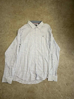Vit rutig skjorta från Tommy Hilfiger - Klassisk vit skjorta från Tommy Hilfiger med ljusblått rutmönster och diskret logga på bröstet. Skjortan har lång ärm, knäppning framtill och button down-krage. Perfekt för en clean och stilren look.