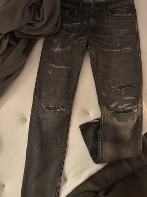 Svarta slitna slim jeans diesel  - Säljer ett par svarta diesel slim jeans med slitningar och distressed detaljer framtill och baktill. Jeansen har fem fickor, bälteshällor och klassisk knappgylf. Perfekta för en edgy streetstil.